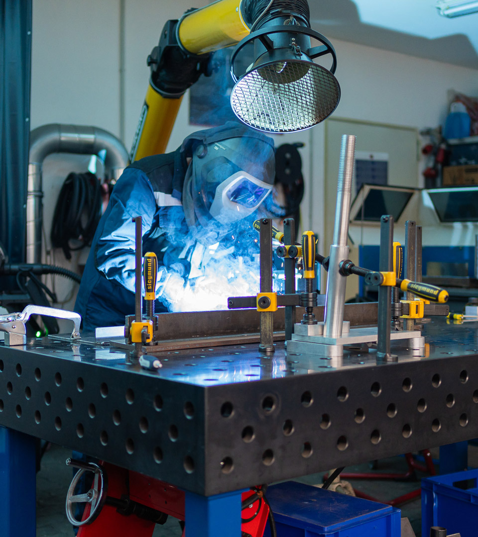 Ein Schweißer arbeitet mit professioneller Schutzausrüstung an einem Spann- und Schweißtisch, während eine Absauganlage den Rauch abführt – industrielle Anwendung bei Betker & König
