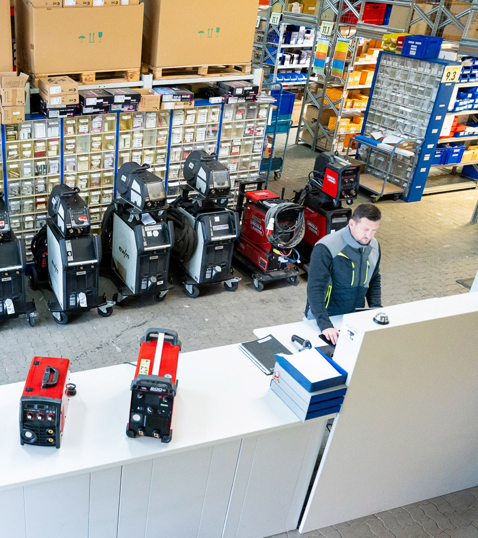 Ein Mitarbeiter bei Betker & König im Lagerbereich, umgeben von Schweißgeräten, Ersatzteilen und Zubehör – Blick auf die Warenausgabe und Sortimentsstruktur